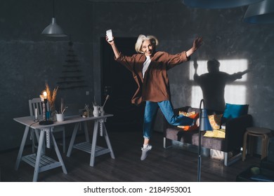 Pleased Caucasian Businesswoman With Smartphone Jumping At Dark Art Studio With Lighting. Small Business And Entrepreneurship. Modern Successful Woman Rejoicing Win And Looking At Camera