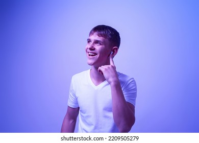 Pleased Caucasian Teenage Boy Pushing On Wireless Earphone For Choosing Music. Blonde Guy Of Zoomer Generation. Modern Youngster Lifestyle. Blue With White Light Background. Studio Shoot. Copy Space