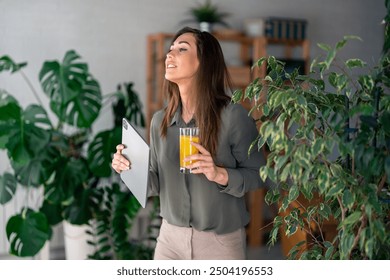 A pleased Caucasian woman sips fresh juice while browsing on her digital tablet, surrounded by lush indoor plants in a cozy home office environment. - Powered by Shutterstock