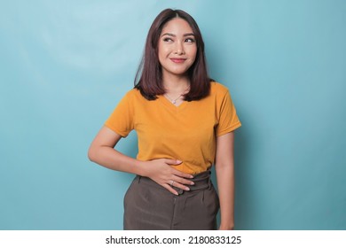 Pleased cheerful Asian woman keeps hand on belly feels full after delicious dinner dressed casually stands thoughtful against blue background. - Shutterstock ID 2180833125