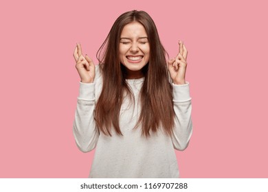 Pleased cheerful brunette woman puts all efforts in wishing good luck, crosses fingers, has faith for better, dressed in oversized sweater, isolated over pink background. People, body language concept