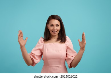 Pleased cheerful smiling european woman with dark hair in pink dress makes big sign with both hands, shapes quite huge object, impressed by size, shapes huge box, isolated on blue background