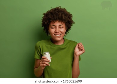 Pleased curly haired woman imagines taste of delicious fresh yoghurt, clenches fist and bites lips awaits for eating healthy product stands with closed eyes against green background. People, nutrition