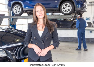 A Pleased Customer, Posing In Front Of Her Justly Fixed Car At A High End Garage, With A Mechanic Tending To Another Vehicle In The Background