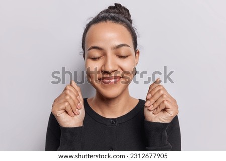 Pleased dark haired Latin woman keeps eyes closed clenches fists dreams about something pleasant smiles and has dimples on cheeks dressed in casual black jumper isolated over white background