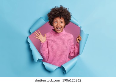 Pleased dark skinned joyful young Afro American woman raises palm dressed in knitted sweater breaks through paper hole of blue background reacts on awesome news. People positive reaction concept