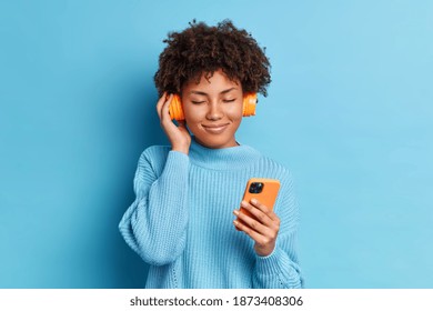 Pleased dark skinned woman with Afro hair holds mobile phone and listens audio track from playlist closes eyes wears jumper isolated over blue background. People technology lifestyle concept - Powered by Shutterstock