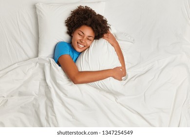 Pleased dark skinned woman enjoys nice dreams during sleeping, embraces pillow, lies in bed comfortably, feels blissful, rests and naps, sleeps well, enjoys fresh soft bedding linen. Top view