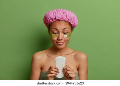 Pleased dark skinned woman holds sanitary napkin applies collagen patches under eyes wrapped in bath towel wears waterproof hat isolated over green background. Hygiene womens health concept.