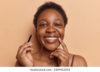 Pleased dark skinned woman and short hair holds lip pencil smiles broadly showcases artistry makeup dressed in t shirt isolated over brown background  Women   beauty procedures concept