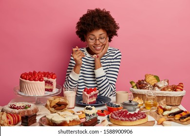 Pleased dark skinned woman tastes yummy strawberry cake, has good appetite, prepared various desserts for festive event, likes eating delicious food containing much calories, isolated on pink