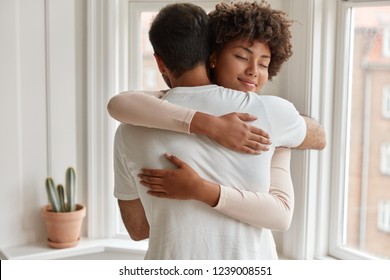 Pleased dark skinned young woman gives warm hug to her boyfriend, being pleased, pose near window, have romantic relationship, stand in cozy room. Husband and wife feel pleased and togetherness