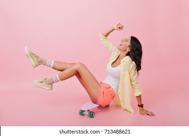 Pleased dark-haired girl in cute shorts and colorful socks playfully posing on skateboard. Indoor portrait of tanned young woman in bright accessories sitting on pink background and laughing. - Powered by Shutterstock