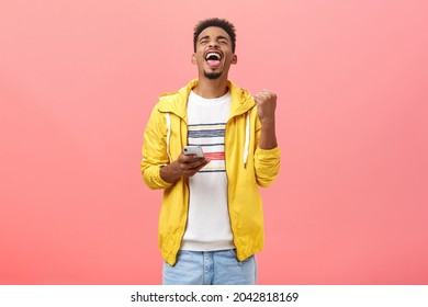 Pleased delighted good-looking african guy triumphing beating score in popular smartphone game raising clenched fist holding cellphone yelling from joy with closed eyes over pink background