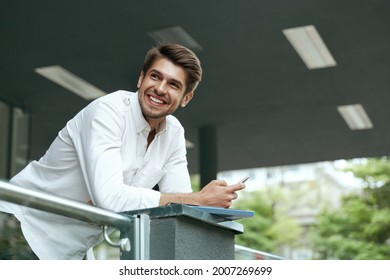 Pleased european businessman holding mobile phone and documents. Young man standing on balcony of modern office building in city. Smiling bearded stylish guy wear white shirt and jeans. Daytime - Shutterstock ID 2007269699