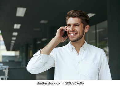 Pleased European Businessman Talking On Mobile Phone. Young Man Standing On Background Of Modern Office Building In City. Smiling Bearded Stylish Guy Wear White Shirt. Daytime