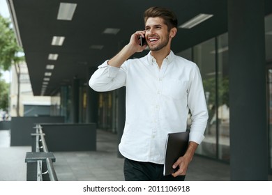 Pleased European Businessman Talking On Mobile Phone And Holding Documents. Young Man Standing On Background Of Modern Office Building In City. Smiling Bearded Guy Wear White Shirt And Jeans. Daytime