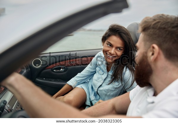 Pleased european couple riding in personal\
cabriolet automobile. Young beautiful woman and adult man looking\
at each other. Concept of trip and journey in car. Idea of freedom.\
Sea background