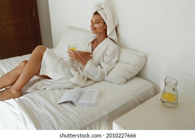 Pleased european girl with closed eyes laying on bed and drink lemonade. Young beautiful woman with under eye patch on face wear bathrobe and wrapped bath towel on head. Concept of rest. Home bedroom - Powered by Shutterstock