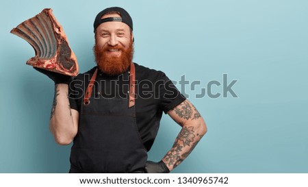Pleased ginger man prepares fresh product for market, carries big piece of meat on rib, involved in packing and distribution, keeps hand on waist, wears black cap and apron. Food technology.