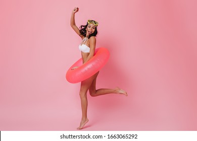 Pleased girl in bikini jumping on pink background. Studio shot of excited young woman with swimming circle.