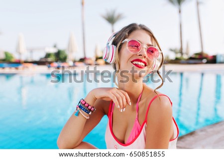 Pleased girl with bright make-up and colorful accessories enjoying southern landscape while listening music. Adorable young woman with bronze skin in white tank-top sitting near pool after swimming