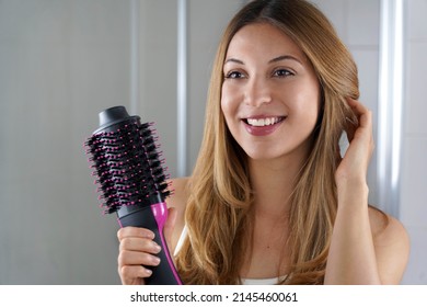Chica encantada sostiene un secador de pelo de cepillo redondo para peinarse en su cuarto de baño en casa. Joven con secador de pelo de un paso y volumizador de salón.