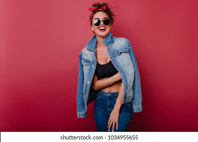 Pleased Girl Wears Jeans Laughing On Claret Background. Spectacular Female Model Looking To Camera Through Black Sunglasses.