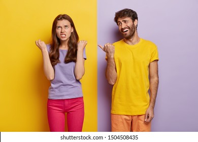 Pleased guy has fun, wears bright yellow t shirt, points thumb at irritated girlfriend who clenches fists with anger, expresses negative emotions, stand in studio against colorful background. Feelings