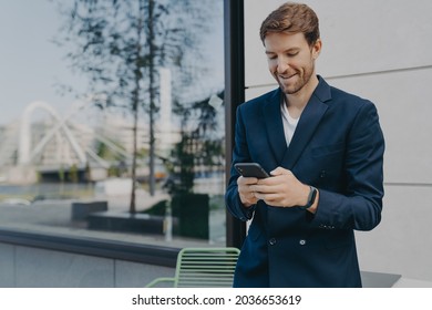 Pleased handsome male CEO or director holds mobile phone waits for call scrolls social networks while waiting for partner near outdoor cafe wastes time online dressed in elegant formal suit. - Powered by Shutterstock