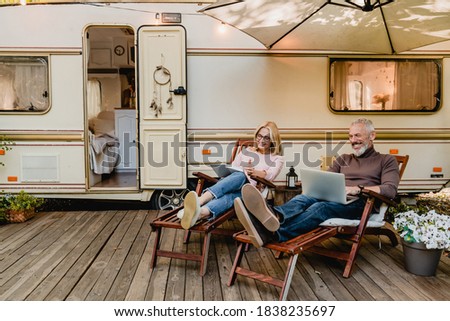 Pleased happy aged couple reading a book and using laptop while relaxing on the deck chairs near the camper van