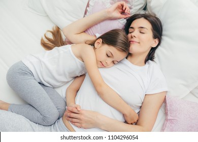 Pleased Little Child Sleeps Near Her Mother, Embraces With Love, Has Pleasant Dreams, Lie On Comfortable Bed. Mum And Cute Daughter Have Good Sleep In Bedroom. Family, Sleeping And Rest Concept