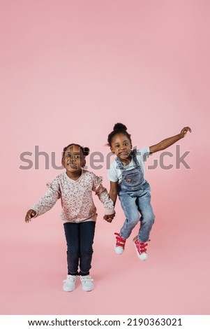 Pleased little girlfriends holding hands and jumping together. African kids sisters spending time together. Childhood concept. Entertainment and leisure. Friendship. Pink background. Copy space.