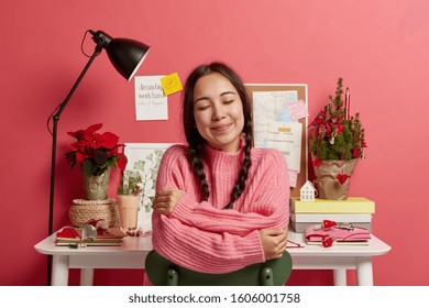 Pleased Lovely Asian Female Embraces Herself, Wears Pink Knitted Sweater, Poses At Cozy Desktop With Decorated Christmas Tree, Eggnog, Desklamp, Spends Holiday In Office, Enjoys Festive Day.