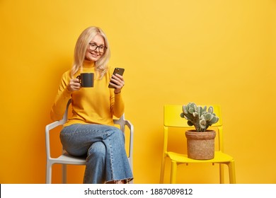 Pleased middle aged woman works distantly from home has coffee break sits on comfortable chair surfs internet via mobie phone has glad expression dressed casually isolated over yellow background - Powered by Shutterstock