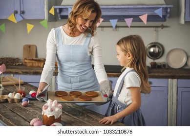 Pleased Mom Teaching Her Daughter Making Easter Sweets. She Is Holding Tray With Cookies While Girl Is Looking At It With Joy