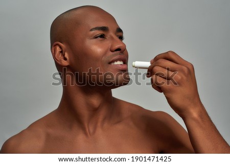 Pleased multiethnic man lifting a tiny tube with lip salve and smearing it over mouth with a smile