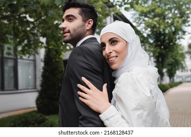 pleased muslim bride in hijab and wedding dress hugging groom
