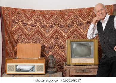 Pleased Old Man In Elegant Clothes Leaned On Retro Tv And Looking At The Camera, Vintage Room With Ornament Wall