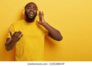 Pleased plump bearded dark skinned man listens romantic song in modern headphones, sings words and gestures with hand, wears yellow t shirt, picks melody for listening on way to work, feels glad - Shutterstock ID 1335288539