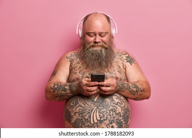 Pleased Plump Man With Naked Tattoed Body, Big Belly, Listens Music In Headphones, Holds Cellular, Downloads Songs In Playlist, Isolated On Pink Background. People, Overweight, Hobby Concept