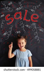 Ein nettes, hübsches kleines Mädchen lächelt und zeigt auf, während es sich gegen eine Tafel mit dem Verkauf Thema Kreide-Aufschriften stellt