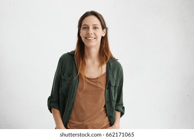 Pleased pretty woman with dyed hair, dark eyes and healthy skin dressed in brown T-shirt, green jacket holding hands in pockets smiling while posing against white concrete wall. People and lifestyle