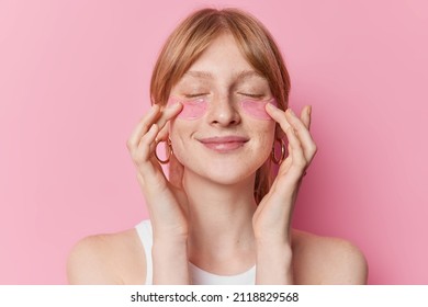 Pleased redhaired female model undergoes beauty procedures appies hydrogel patches under eyes to remove puffiness and fine lines wears casual t shirt isolated over pink background. Skin care - Powered by Shutterstock