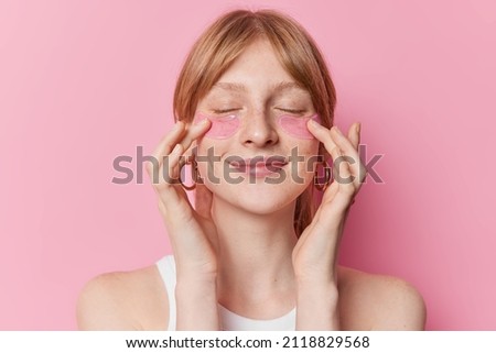 Pleased redhaired female model undergoes beauty procedures appies hydrogel patches under eyes to remove puffiness and fine lines wears casual t shirt isolated over pink background. Skin care