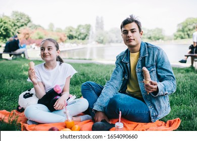 Pleased relaxed teen guy with little sister having rest on picnic sitting on orange plaid on green grass in park and eating ice cream