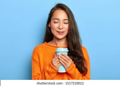 Pleased restful girl with Asian appearance, keeps eyes closed, smiles gently, enjoys drinking aromatic espresso from takeout cup, wears orange jumper, isolated over blue background. People and drink