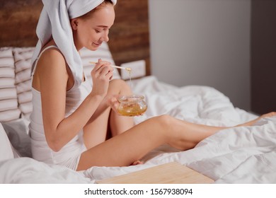 Pleased romantic girl sitting on white sheet with soft bath towel on head, mixing liquid paste for depilation, time for making beautiful skin, removing hairs using wax - Shutterstock ID 1567938094