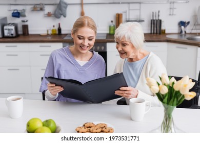 Pleased Senior Woman And Nurse Looking At Photo Album In Kitchen