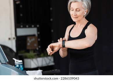 pleased senior woman with wireless earphone checking activity on fitness tracker in gym - Powered by Shutterstock